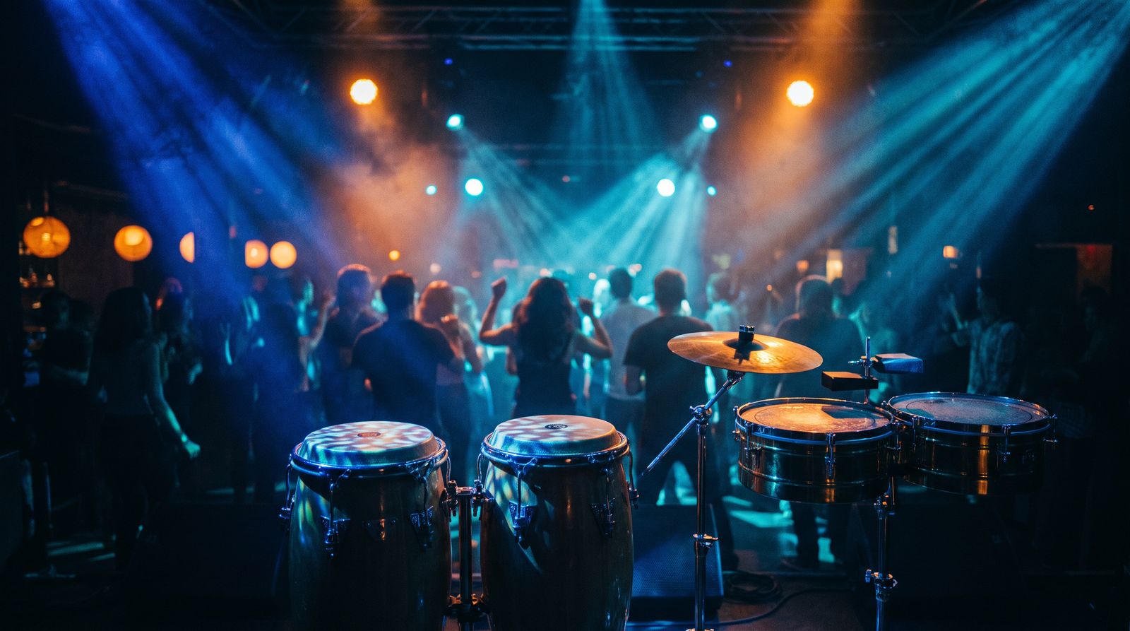 Congas y timbales sobre la tarima, con el público bailando al fondo bajo luces azules y naranja.